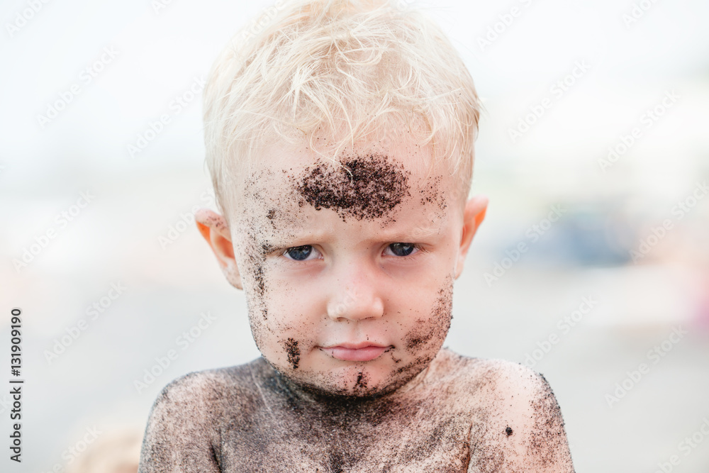 Funny photo of happy baby boy on beach with dirty face covered with black sand. Family travel, healt