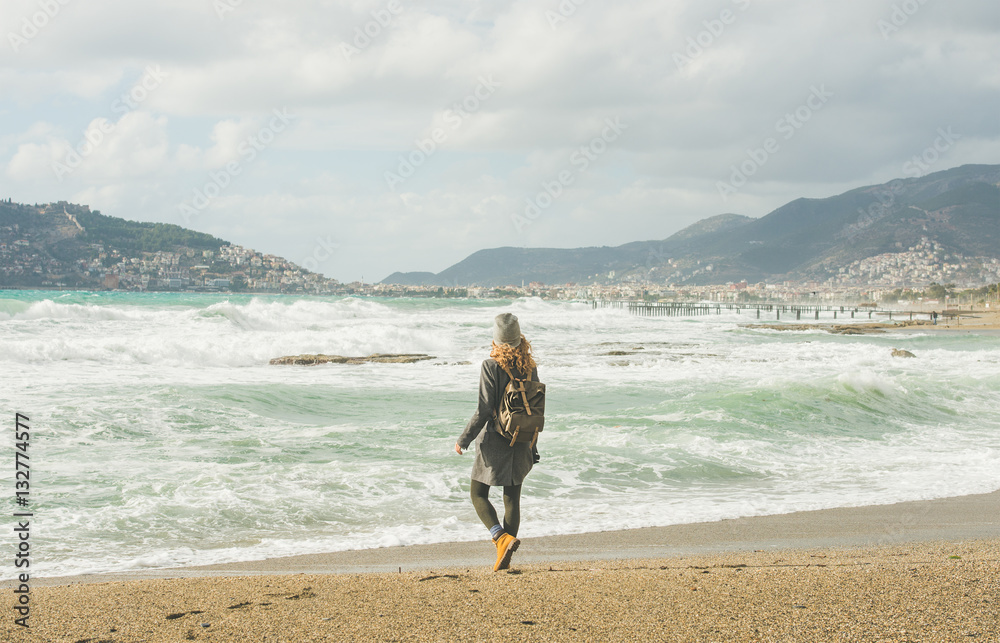 Young woman traveler walking on sandy beach and looking at stormy Mediterranean sea in winter in Ala