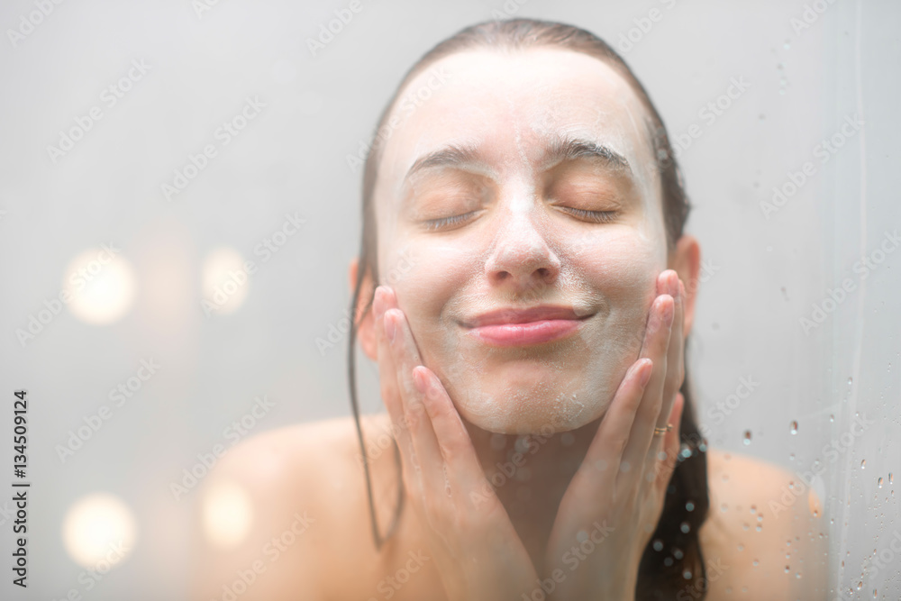 Close-up portrait of a woman with soap on her wet face standing behind the glass in the shower. Imag