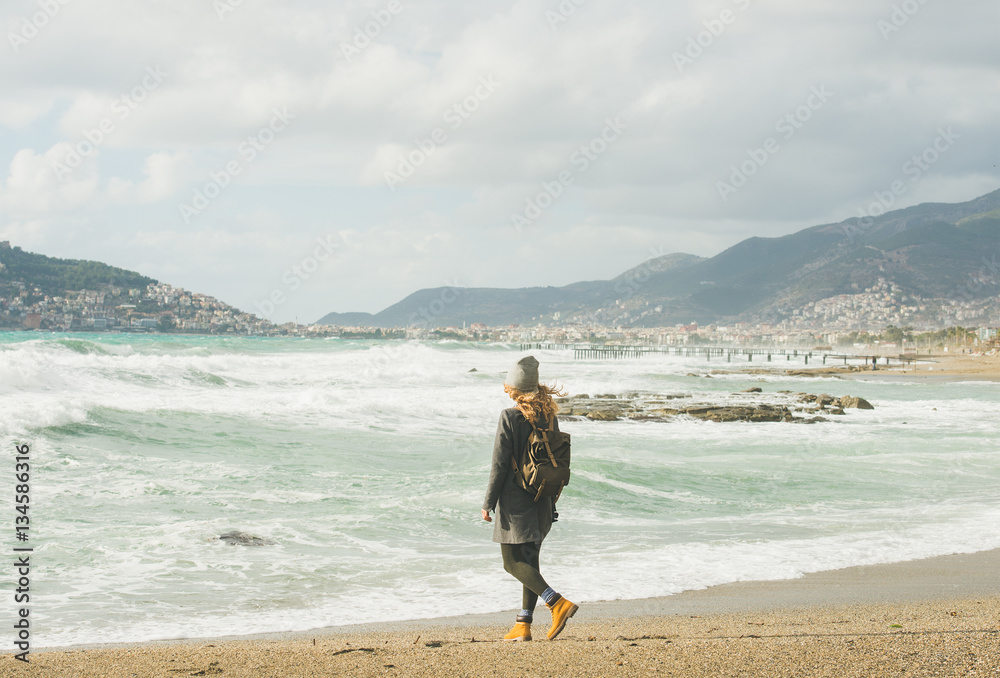 背着背包，穿着灰色外套的年轻女性旅行者在沙滩上行走，看着暴风雨的Mediter