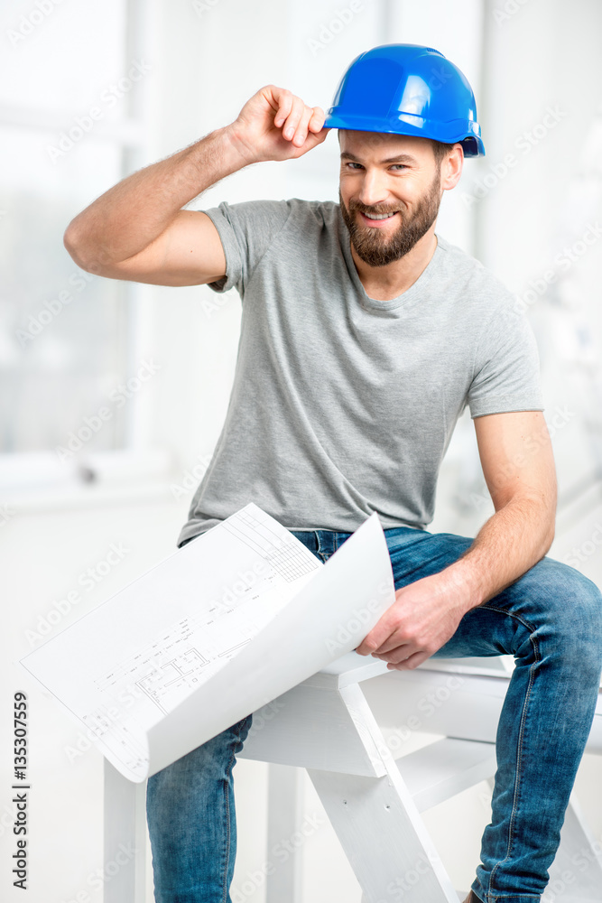 Portrait of a handsome builder, foreman or repairman in the helmet sitting with drawings on ladder i
