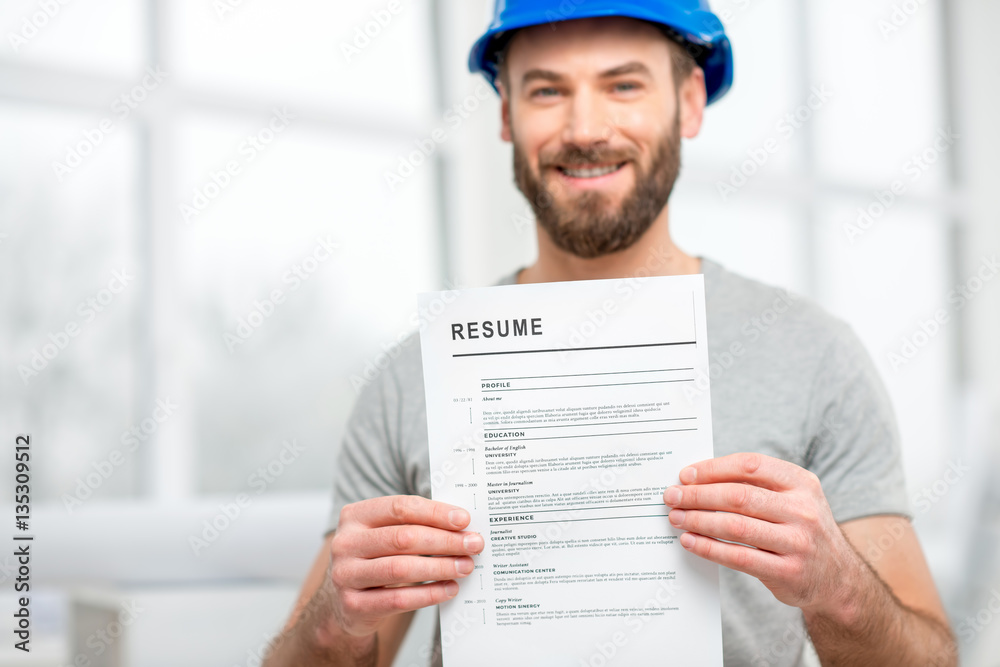 Handsome builder or worker in protective helmet holding his resume standing in the white interior. I