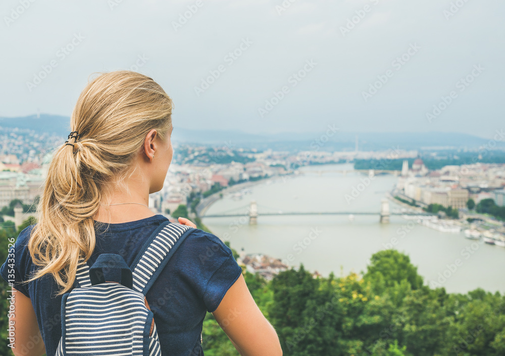 Young blond woman traveler with backpack looking at Danube and Budapest river from hill, rear view, 