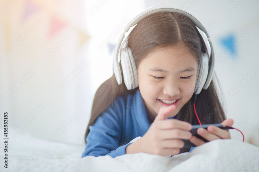 Pretty Asian girl using headphone for listen music by smartphone on the bed in her decorated bedroom