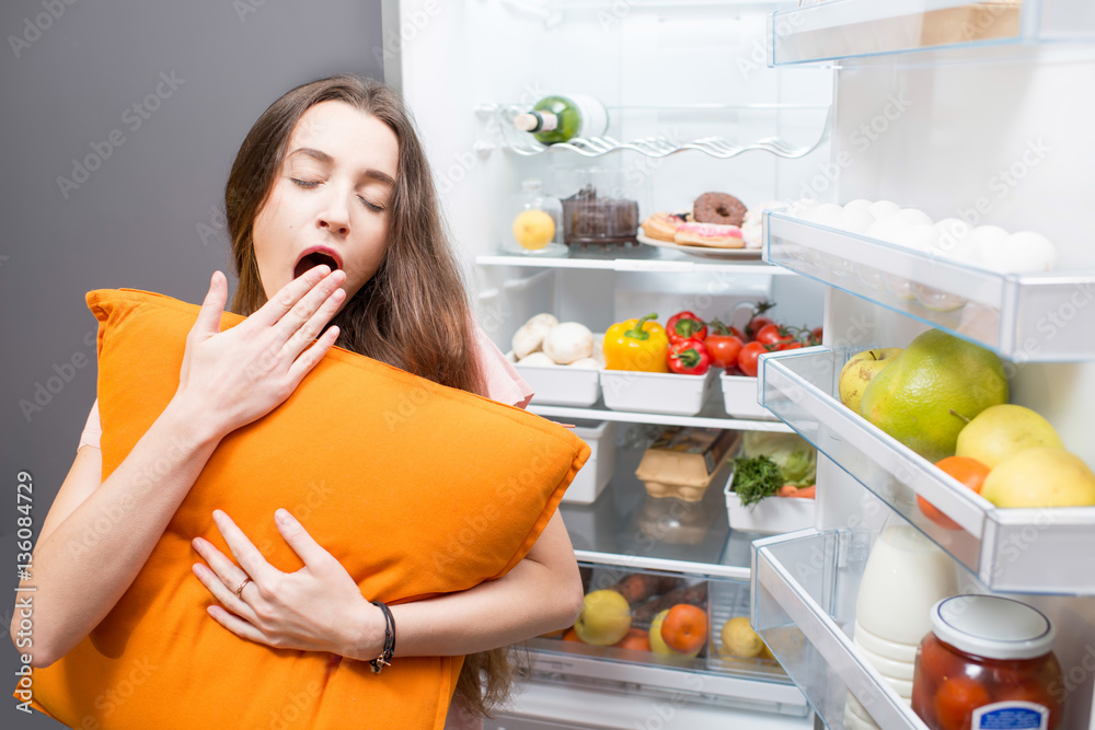 Young woman with pillow yawning standing in front of the refrigerator. Concept of eating during the 