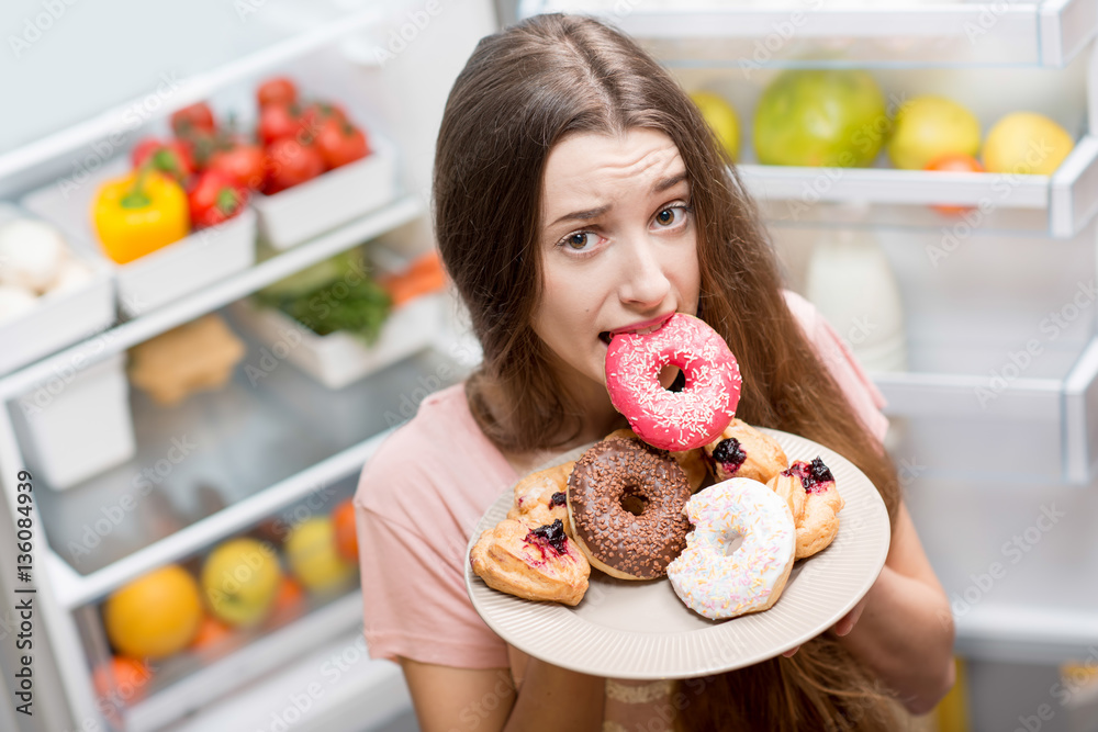 Young woman in the sleepwear eating sweet donuts near the refrigerator