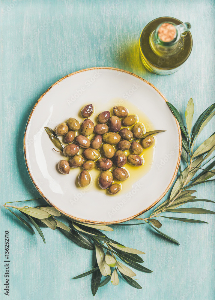 Pickled green Mediterranean olives on white ceramic plate, olive tree branch and virgin olive oil in