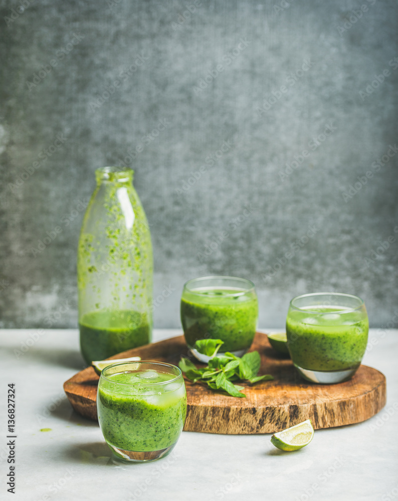 Fresh smoothie in bottle and glasses with ice cubes, mint, lime on wooden board, grey concrete wall 