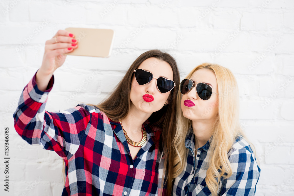 Female friends in checkered shirts making selfie portrait with phone on the white wall background