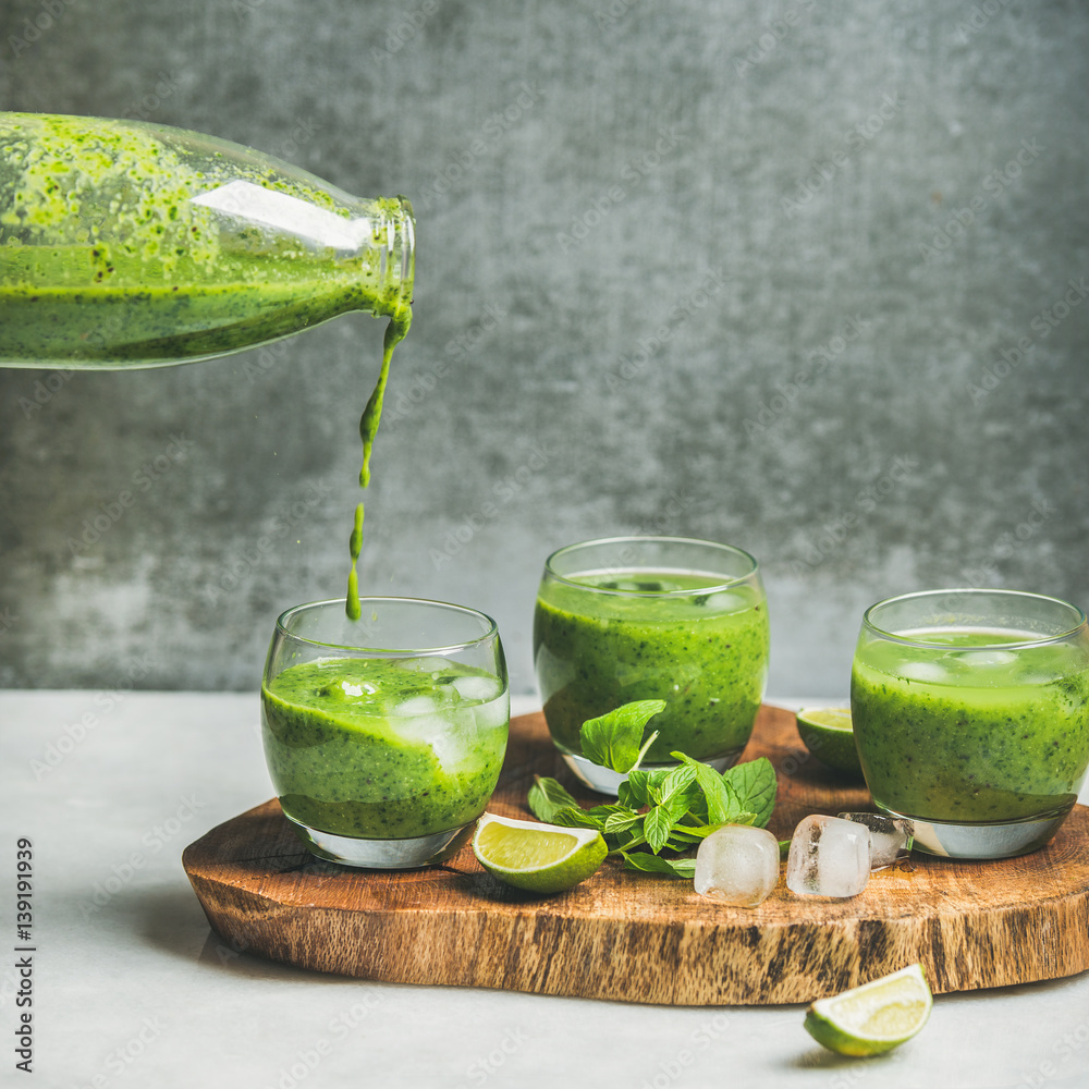 Fresh smoothie in glasses pouring from bottle with ice cubes, mint, lime on wooden board, grey concr