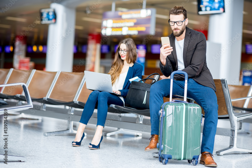 Elegant business couple sitting with laptop, phone and suitcase at the waiting hall in the airport. 