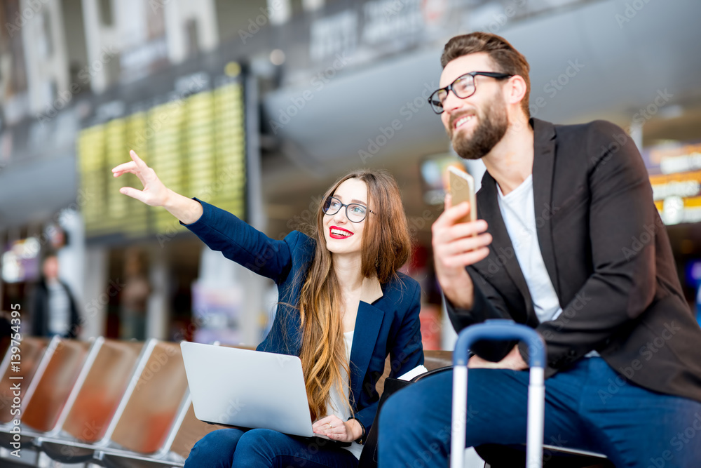 一对优雅的商务情侣坐在机场候机厅，手里拿着笔记本电脑、手机和行李箱。