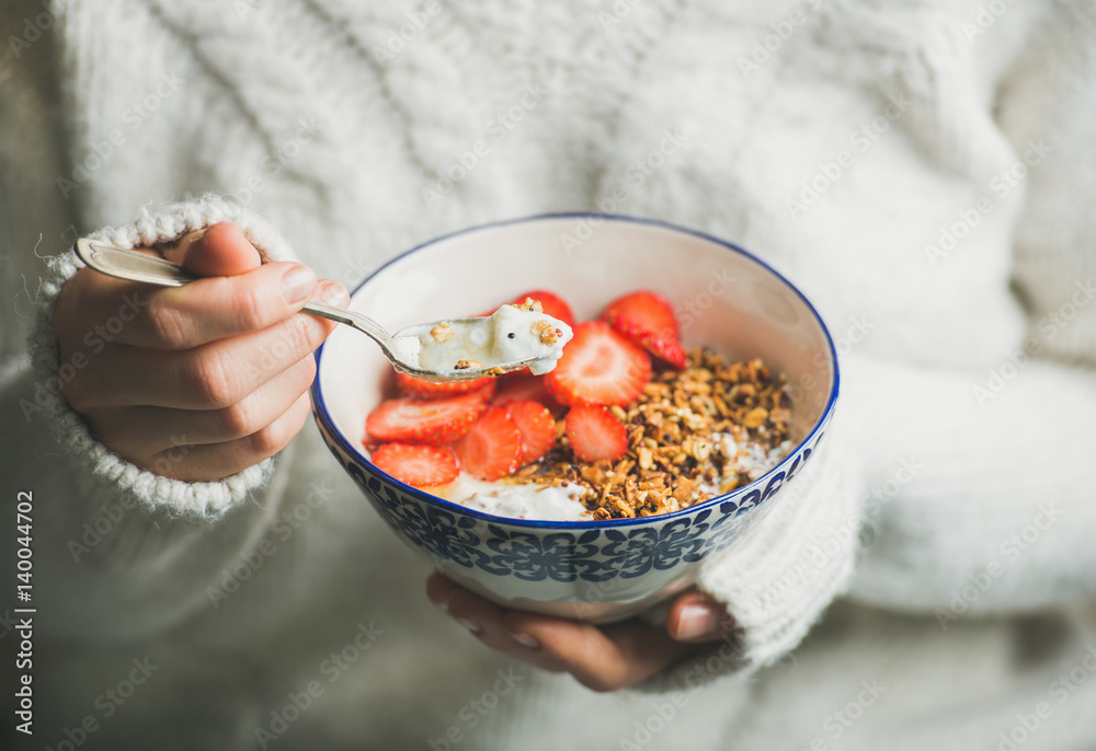 Healthy breakfast greek yogurt, granola and strawberry bowl in hands of woman wearing white loose kn