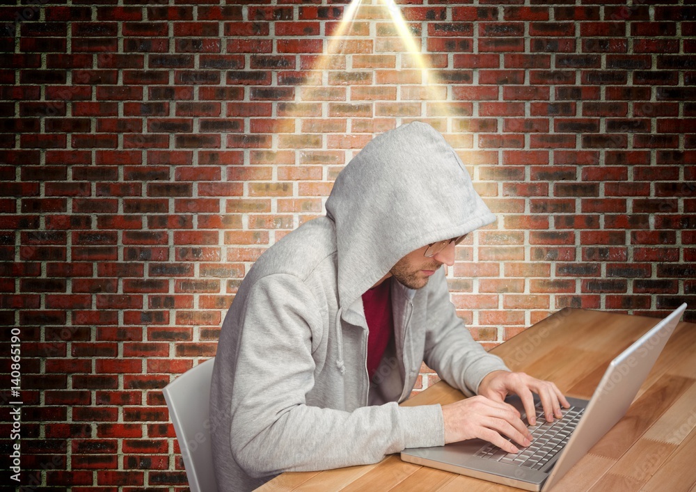 一名身穿连帽上衣的男子靠着砖墙坐着，正在使用笔记本电脑