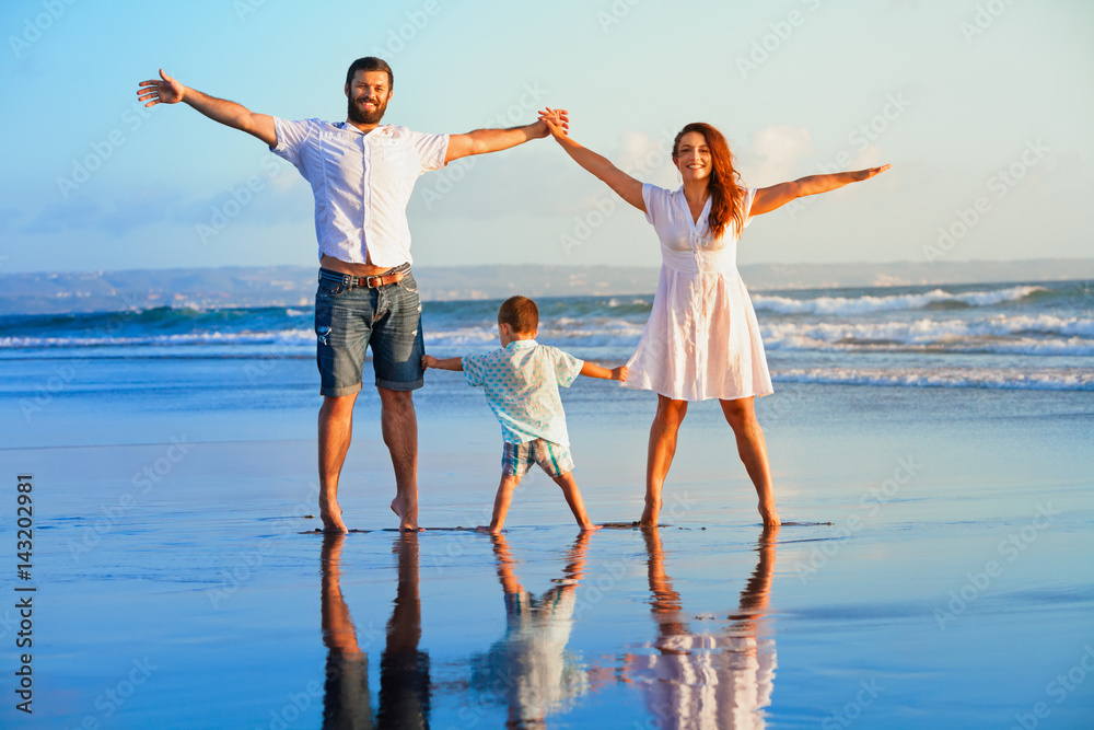 Happy family - father, mother, baby son hold hands, walk and jump together by water pool on sunset b