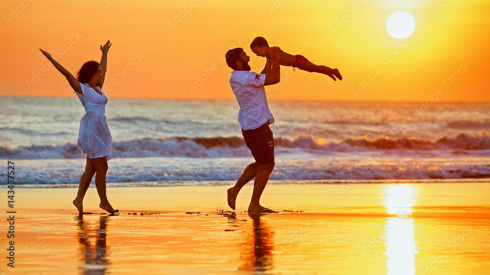 幸福的家庭——父亲、母亲、宝贝儿子沿着日落的边缘在黑沙滩上冲浪，玩得很开心