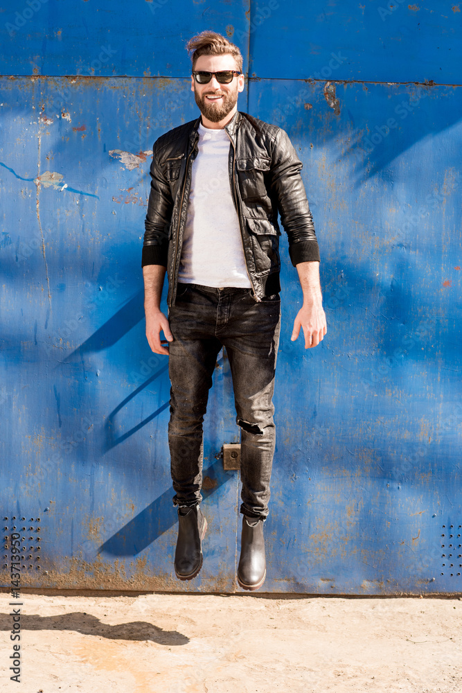 Portrait of a handsome jumping man dressed in white t-shirt, jacket and jeans on the blue rusty wall
