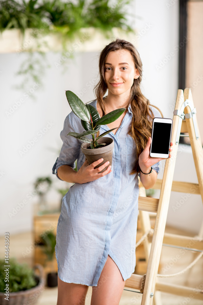 年轻女子站在梯子上，拿着电话和花盆，在家里种绿色植物