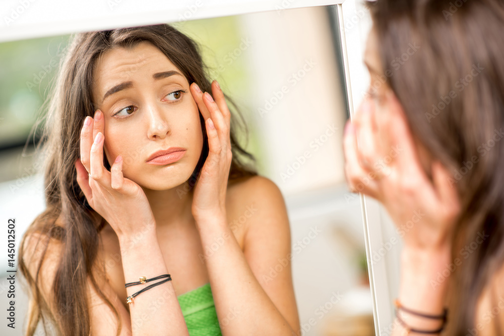 一名年轻女子站在浴室里用毛巾站着，看着镜子，脸上被拧成一团
