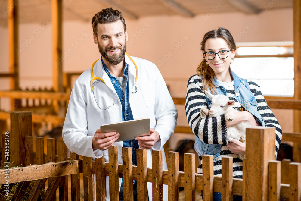 Handsome veterinarian in medical gown and young woman taking care about the baby goat standing indoo