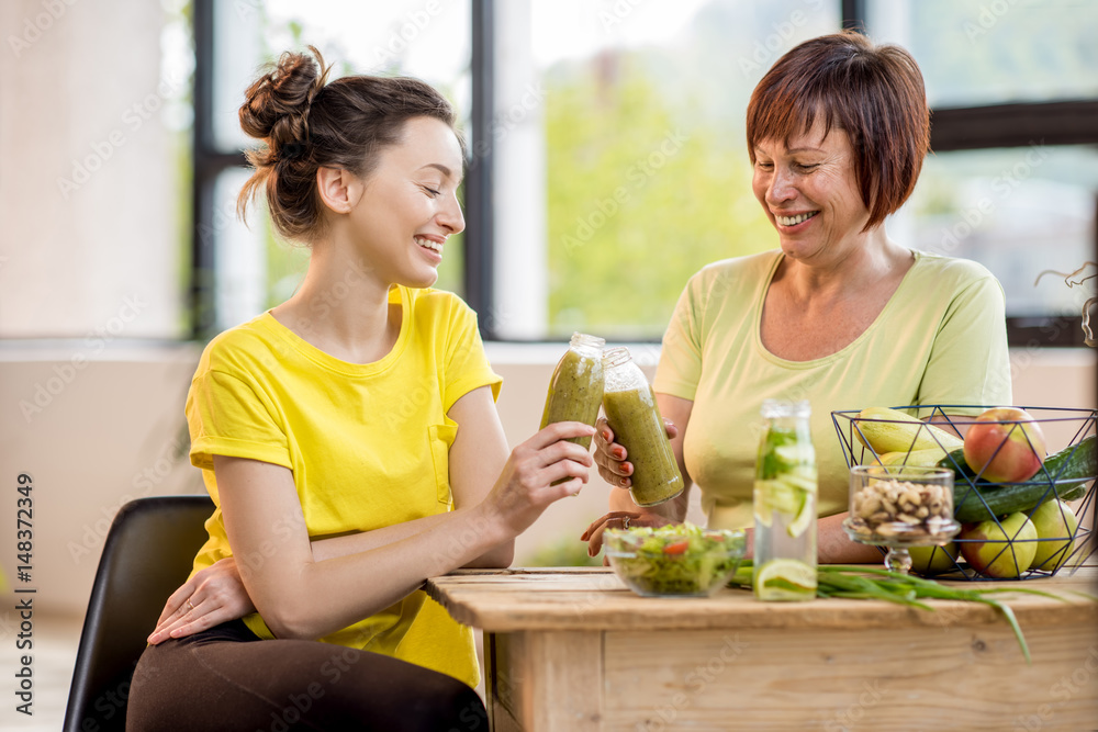 Young and older women sitting with healthy food and fresh drinks after the sports training indoors o
