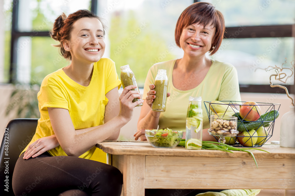 Young and older women sitting with healthy food and fresh drinks after the sports training indoors o