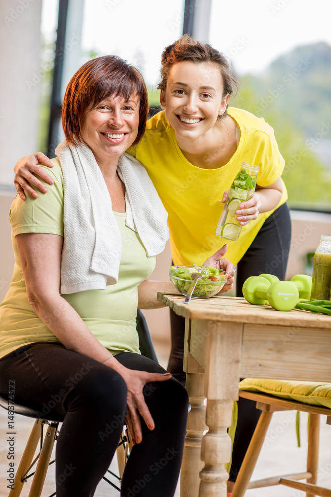 Young and older women exercising with dumbbells and eating healthy food indoors on the window backgr