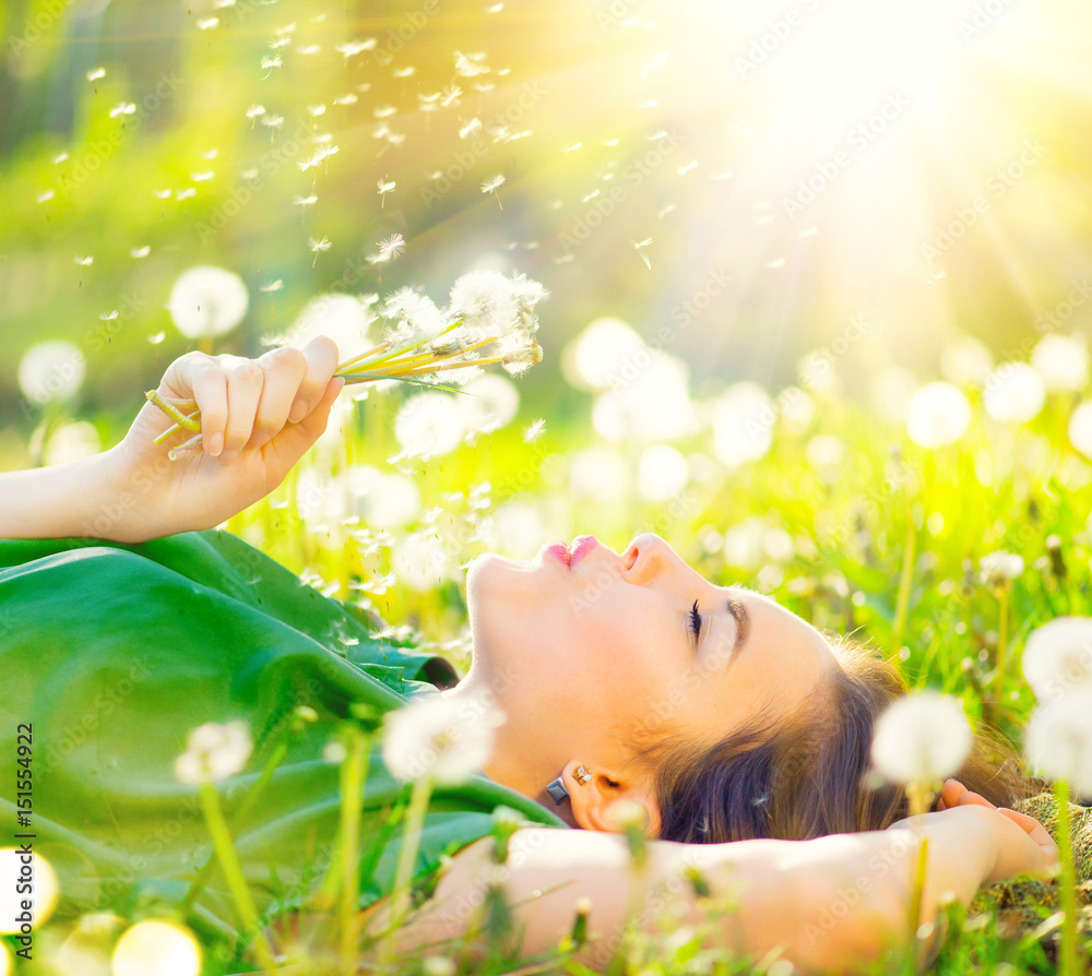 美丽的年轻女子躺在绿草中的田野上，吹着蒲公英