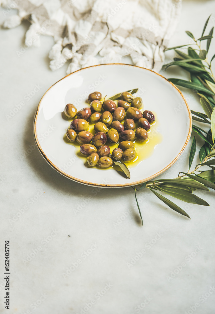 在白色陶瓷盘子和橄榄树枝上用初榨橄榄油腌制的绿色地中海橄榄