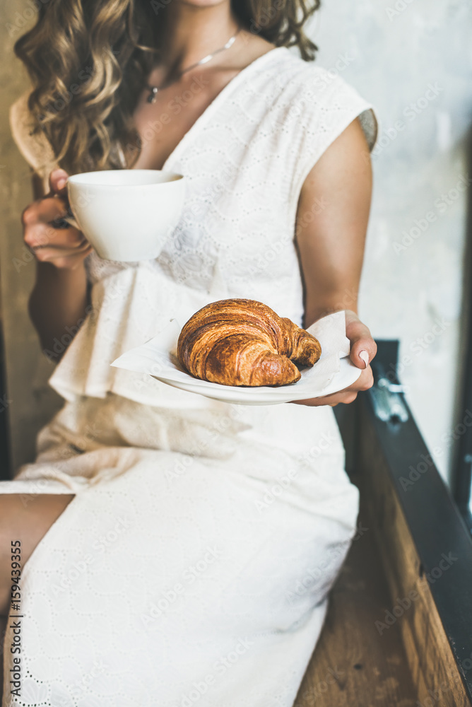 法国或意大利早餐。年轻的金发女子穿着白色连衣裙，手里拿着新鲜的羊角面包。