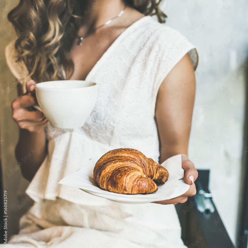 法国或意大利早餐。年轻的金发女子穿着白色连衣裙，手里拿着新鲜的羊角面包。