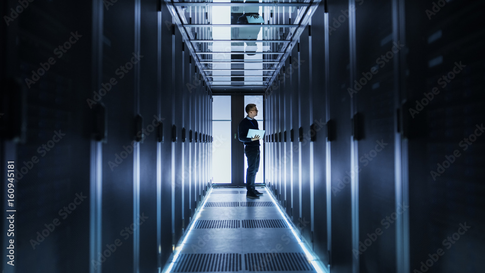 Male IT Engineer Works on a Laptop at the end of a Corridor in a Big Data Center. Rows of Rack Serve