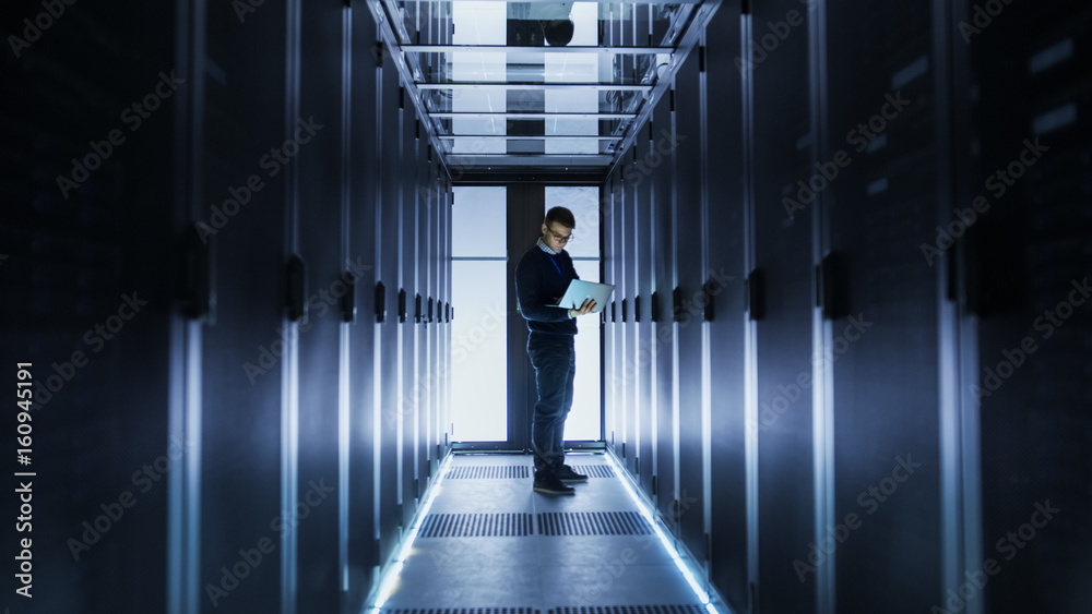 男IT工程师在大数据中心走廊尽头的笔记本电脑上工作。一排排机架服务