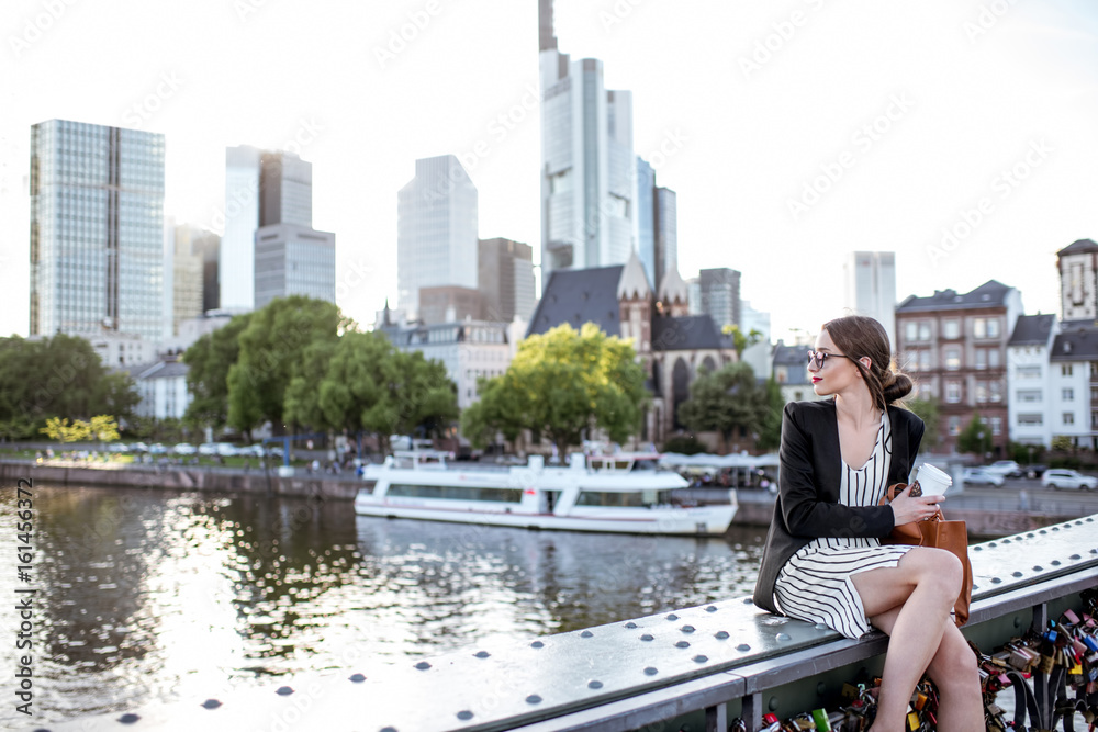 年轻的女商人坐在法兰克福市的桥上，在户外喝咖啡休息