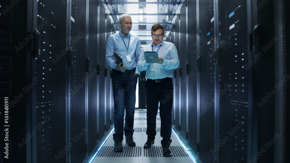 在数据中心，两名IT工程师走过一排排服务器机架。他们在平板电脑上工作