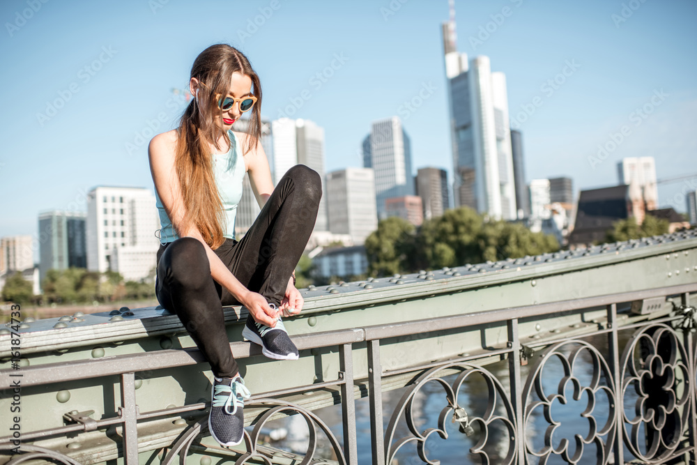 Young woman lacing up sports shoes sitting on the bridge during the training with great view on the 