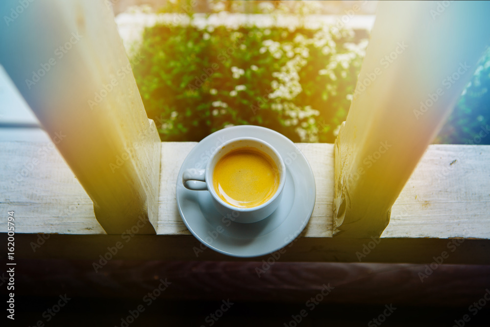 A cup of white on a saucer of morning coffee in the sun on a glass table. The steam of the morning s
