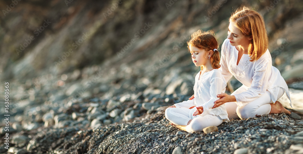 快乐的家庭母亲和孩子做瑜伽，在海滩上以莲花姿势冥想