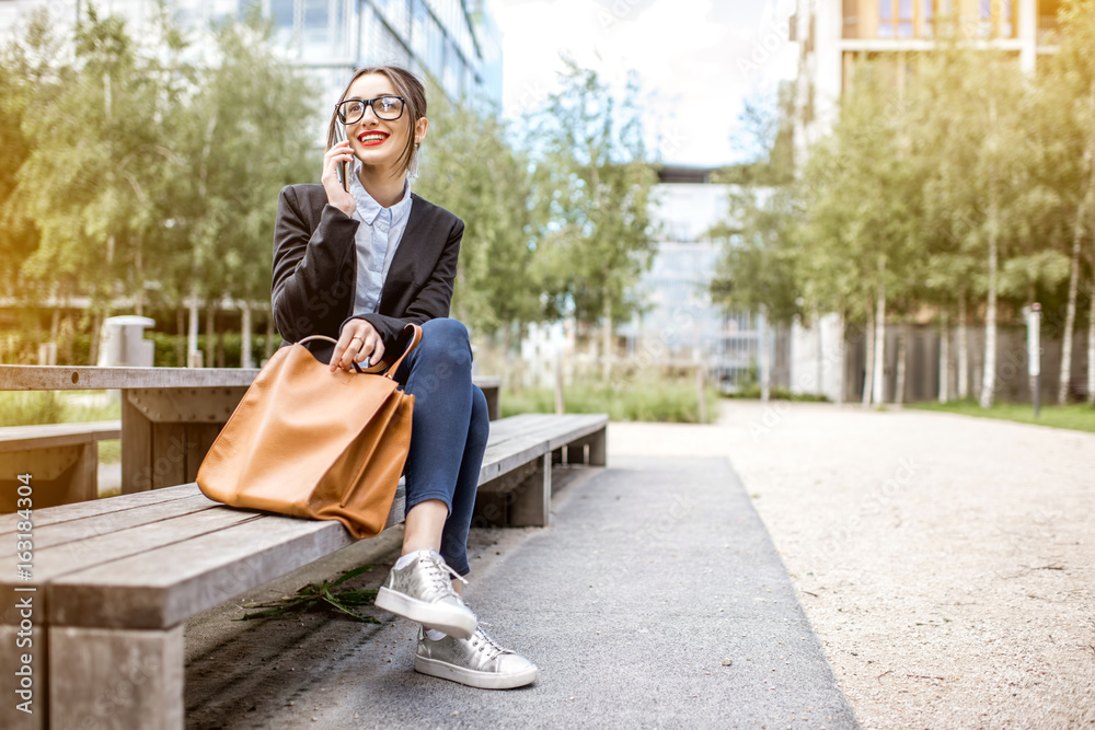 Young businesswoman sitting on the bench with phone outdoors at the modern office district