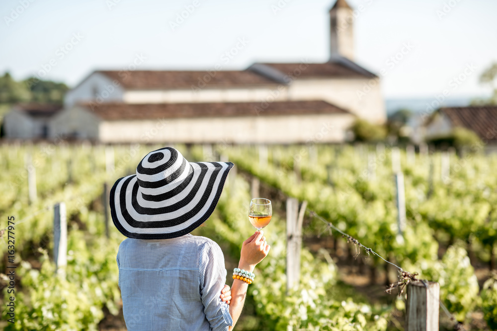 Young woman with wine glass standing on the beautiful vineyard during the sunset