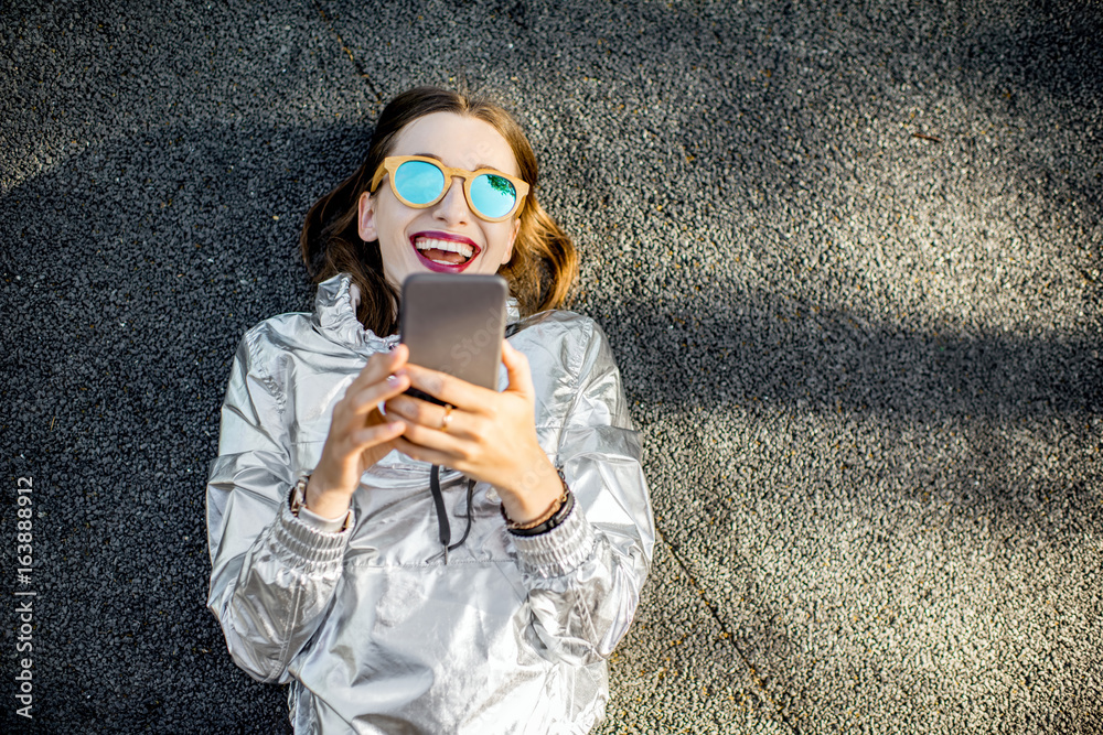 一位穿着银色夹克、拿着手机躺在户外柏油路上的现代女性的生活方式画像