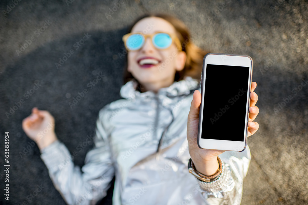 Lifestyle portrait of a modern woman in silver jacket showing phone with blank screen lying outdoors