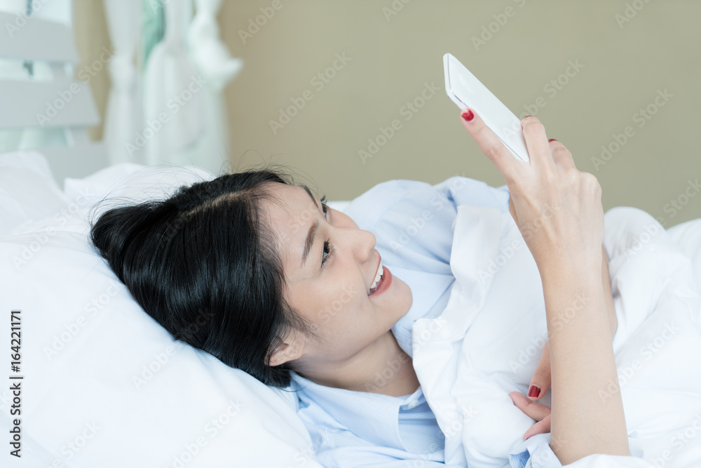 美丽的亚洲少女躺在白色床上，在家里的卧室里用手机。Technol