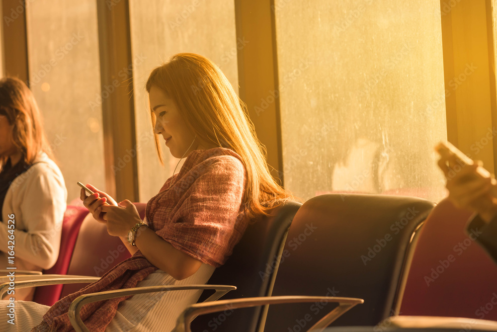 年轻美女拿着旅行袋坐在机场，在机场使用带耳机的智能手机等待f