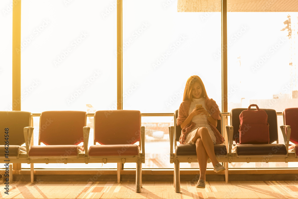 年轻美女拿着旅行袋坐在机场，在机场使用带耳机的智能手机等待f