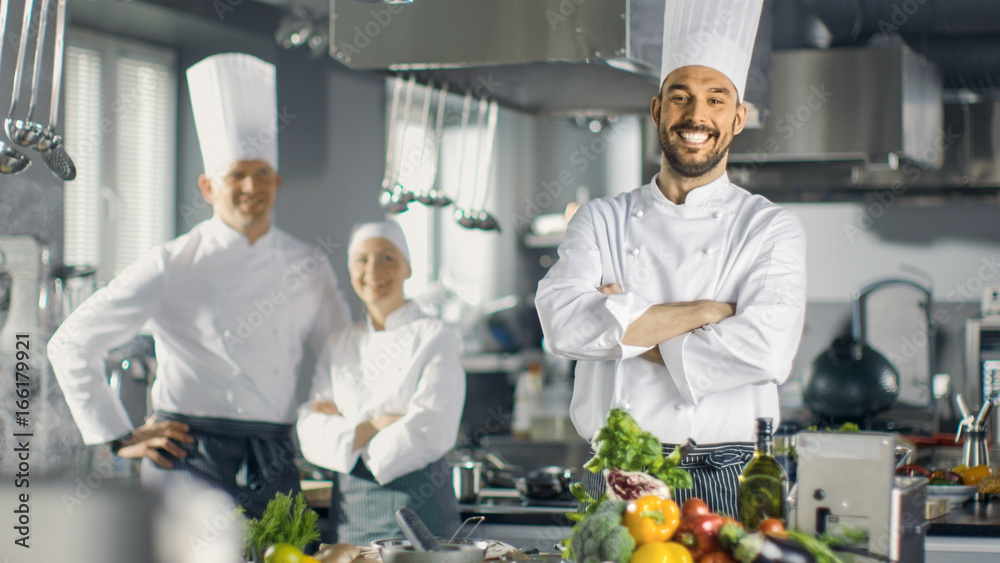 Famous Chef of a Big Restaurant Crosses Arms and Smiles in a Modern Kitchen. His Staff in Smiling in