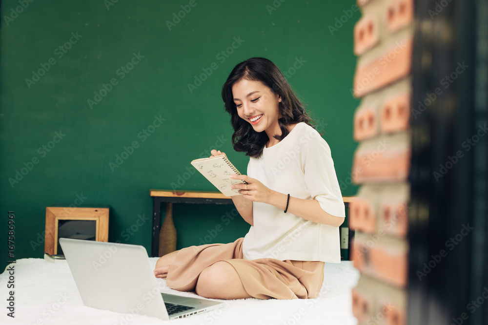 美丽的年轻微笑的女人坐在家里的床上用笔记本电脑工作