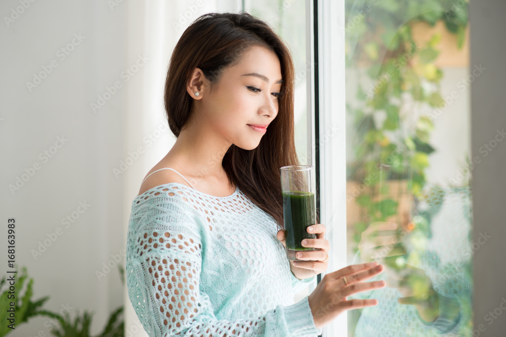 Asian woman drinking green fresh vegetable juice from glass at home