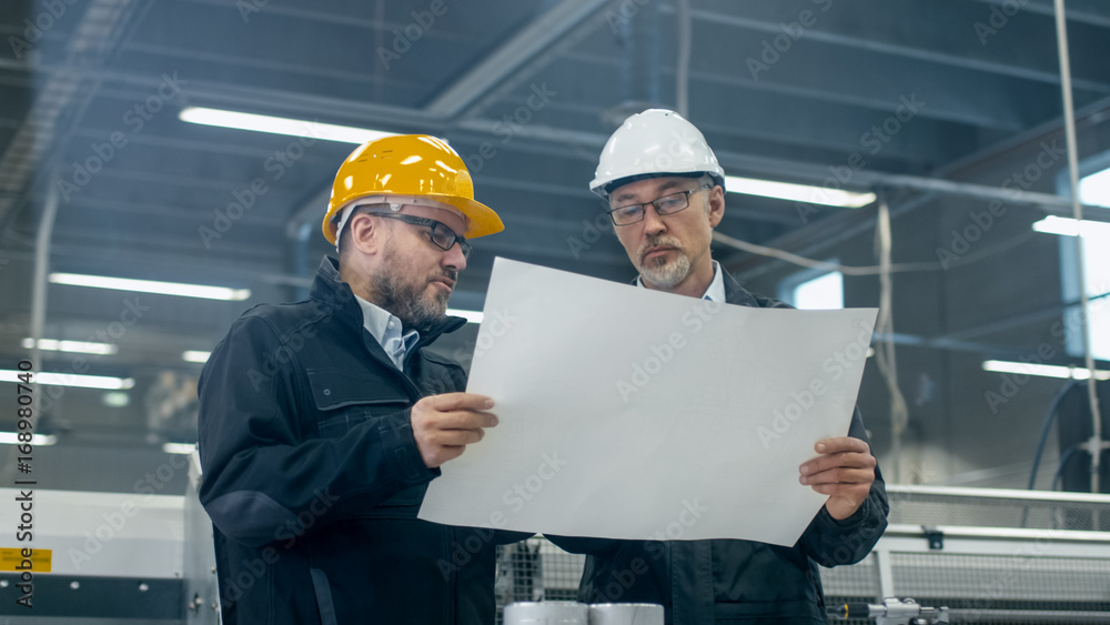 两名戴着安全帽的工程师站在工厂里讨论蓝图。