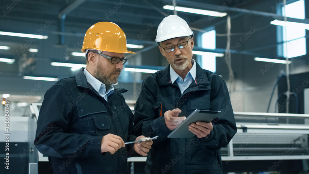 两名戴着安全帽的工程师站在工厂里讨论平板电脑上的信息。